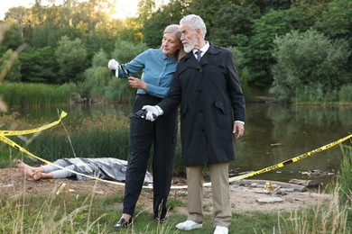Photo of Forensic expertise. Detectives working at crime scene with dead man's body near lake outdoors