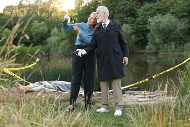 Photo of Forensic expertise. Detectives working at crime scene with dead man's body near lake outdoors