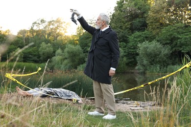 Photo of Forensic expertise. Detective with evidence working at crime scene with dead man's body near lake outdoors