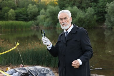 Photo of Forensic expertise. Detective with magnifying glass working at crime scene with dead man's body near lake outdoors