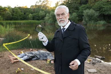 Photo of Forensic expertise. Detective with magnifying glass working at crime scene with dead man's body near lake outdoors