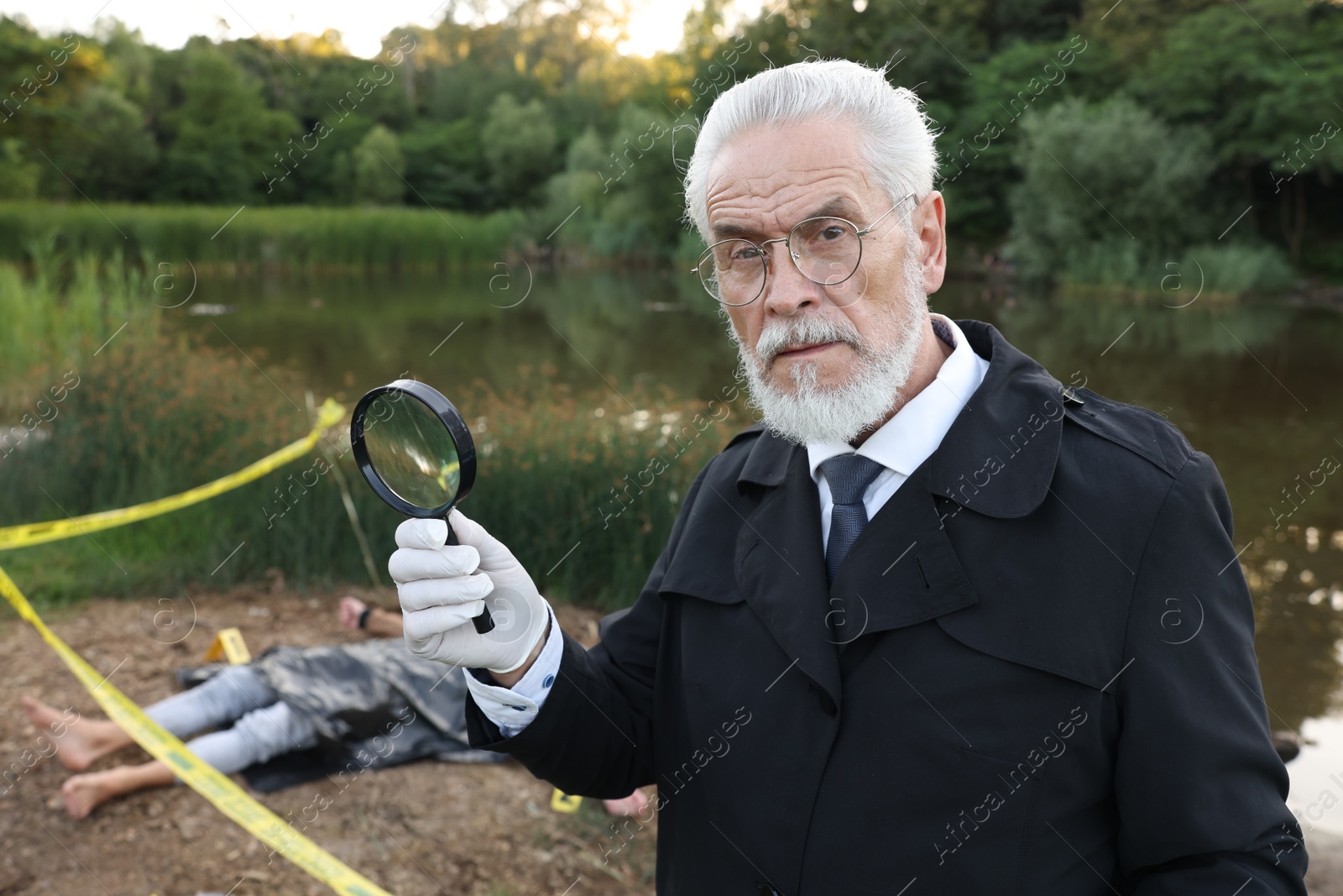 Photo of Forensic expertise. Detective with magnifying glass working at crime scene with dead man's body near lake outdoors