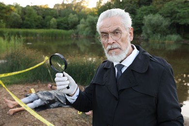 Photo of Forensic expertise. Detective with magnifying glass working at crime scene with dead man's body near lake outdoors