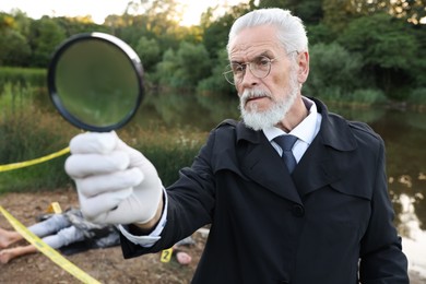 Photo of Forensic expertise. Detective with magnifying glass working at crime scene with dead man's body near lake outdoors