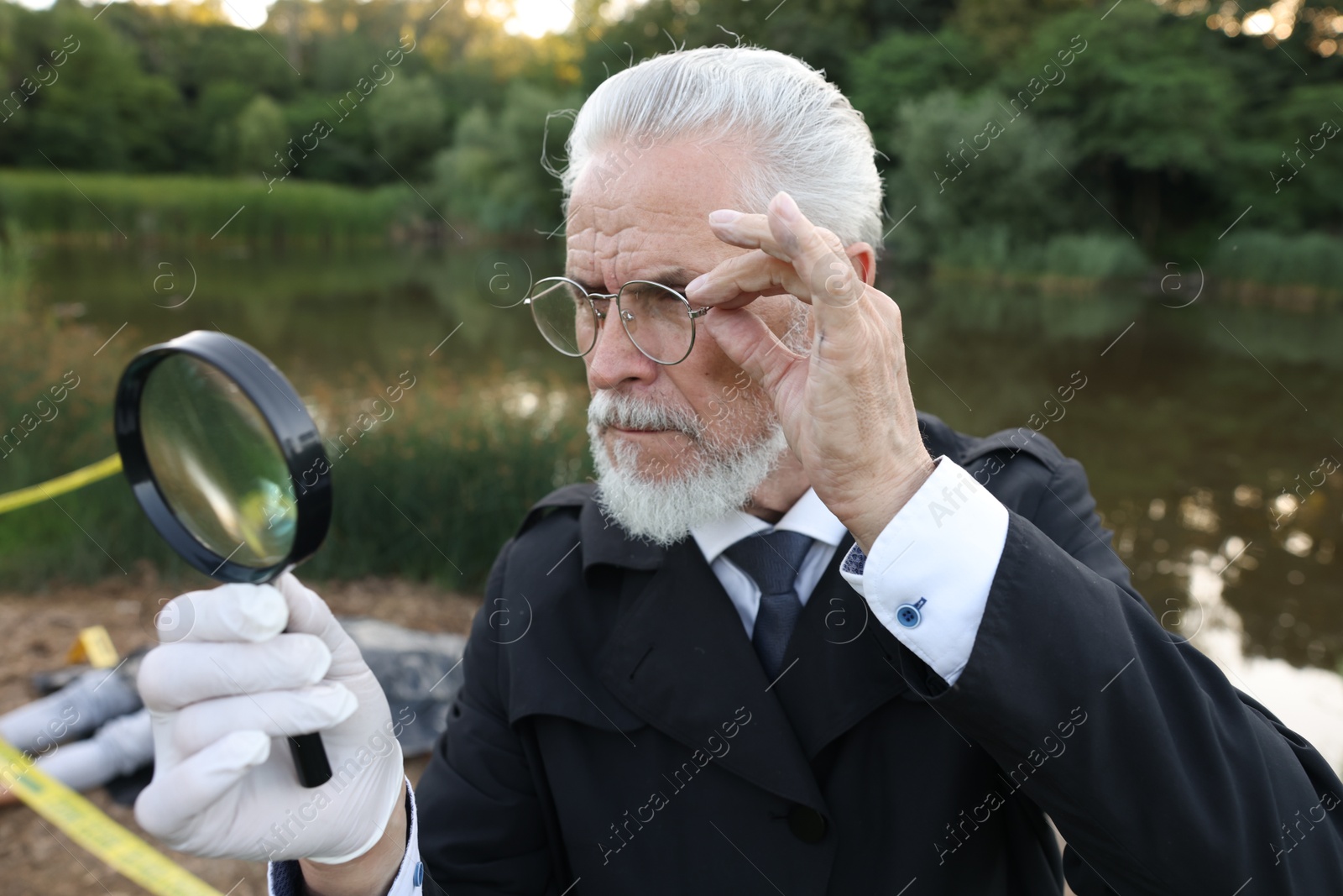 Photo of Forensic expertise. Detective with magnifying glass working at crime scene with dead man's body near lake outdoors