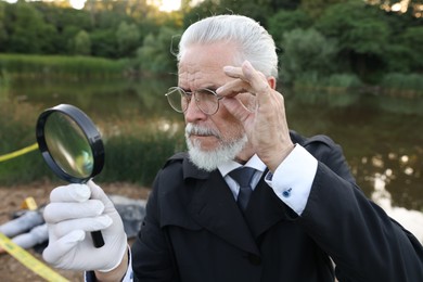 Photo of Forensic expertise. Detective with magnifying glass working at crime scene with dead man's body near lake outdoors