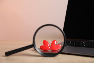 Photo of Online dating site. Laptop, red hearts and magnifying glass on wooden table, closeup