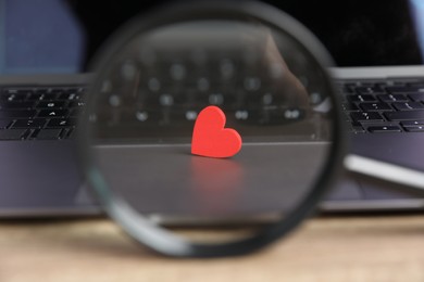 Photo of Online dating site. Laptop, red heart and magnifying glass on table, closeup