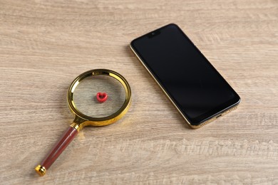 Photo of Online dating site. Smartphone, red heart and magnifying glass on wooden table, above view