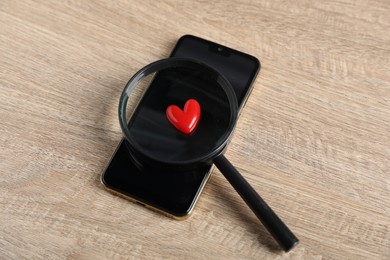Photo of Online dating site. Smartphone, red heart and magnifying glass on wooden table, above view