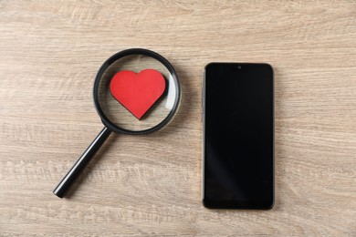 Photo of Online dating site. Smartphone, red heart and magnifying glass on wooden table, flat lay