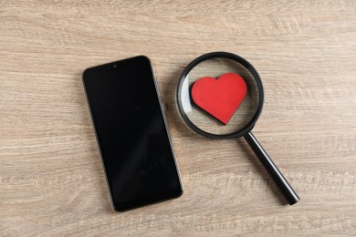 Photo of Online dating site. Smartphone, red heart and magnifying glass on wooden table, flat lay