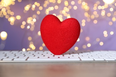 Photo of Online dating site. Red heart and computer keyboard on table against blurred lights, closeup. Bokeh effect