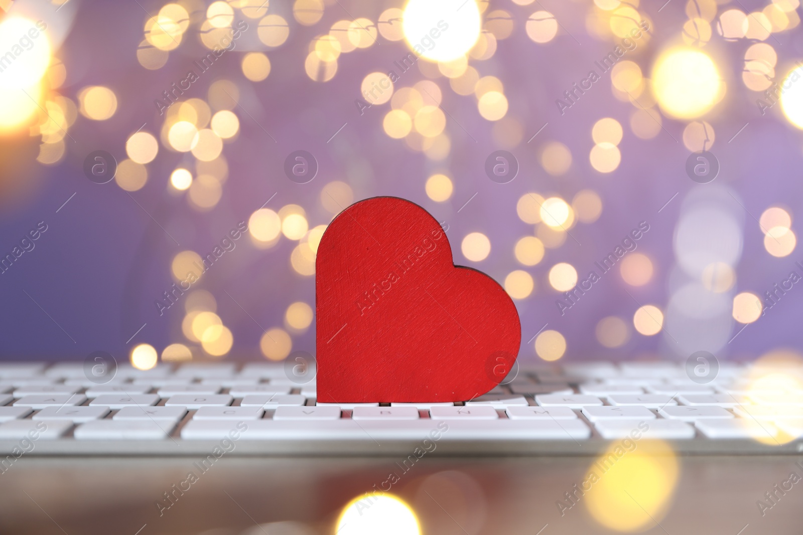 Photo of Online dating site. Red heart and computer keyboard on table against blurred lights, closeup. Bokeh effect