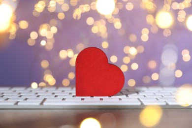 Photo of Online dating site. Red heart and computer keyboard on table against blurred lights, closeup. Bokeh effect