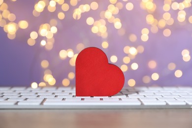 Photo of Online dating site. Red heart and computer keyboard on table against blurred lights, closeup. Bokeh effect