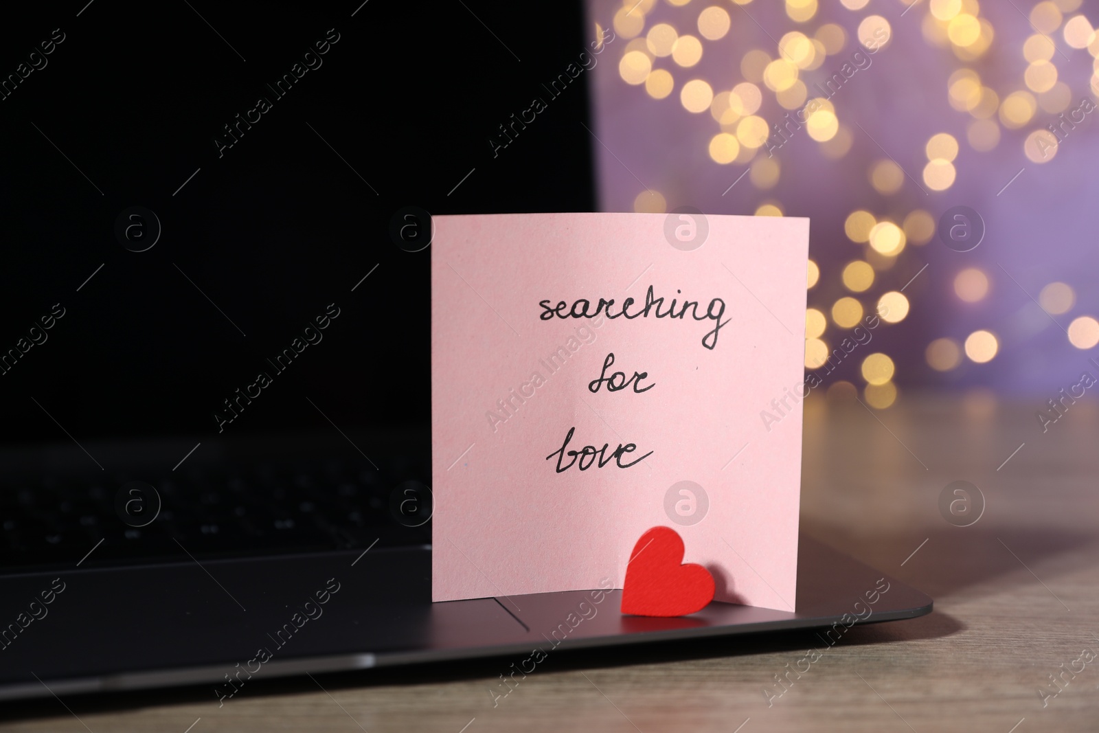Photo of Online dating site. Note with text Searching For Love, red heart and laptop on wooden table against blurred lights, closeup. Bokeh effect