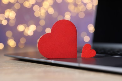Photo of Online dating site. Red hearts and laptop on wooden table against blurred lights, closeup. Bokeh effect