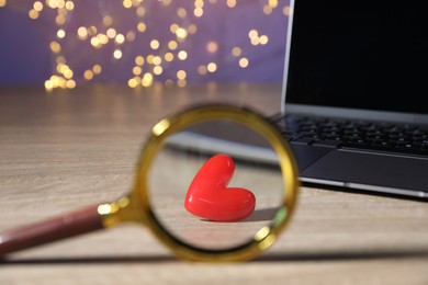 Photo of Online dating site. Red heart, magnifying glass and laptop on wooden table against blurred lights, closeup. Bokeh effect
