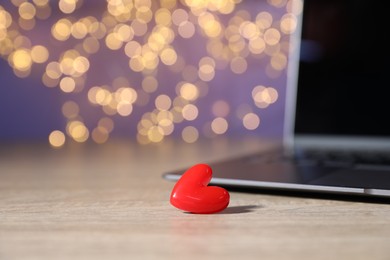 Photo of Online dating site. Red heart and laptop on wooden table against blurred lights, closeup. Bokeh effect