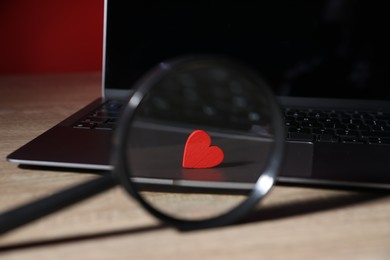 Photo of Online dating site. Red heart, magnifying glass and laptop on wooden table, closeup
