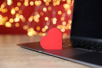 Photo of Online dating site. Red heart and laptop on wooden table against blurred lights, closeup. Bokeh effect