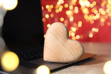 Photo of Online dating site. Wooden heart and laptop on table against blurred lights, closeup. Bokeh effect