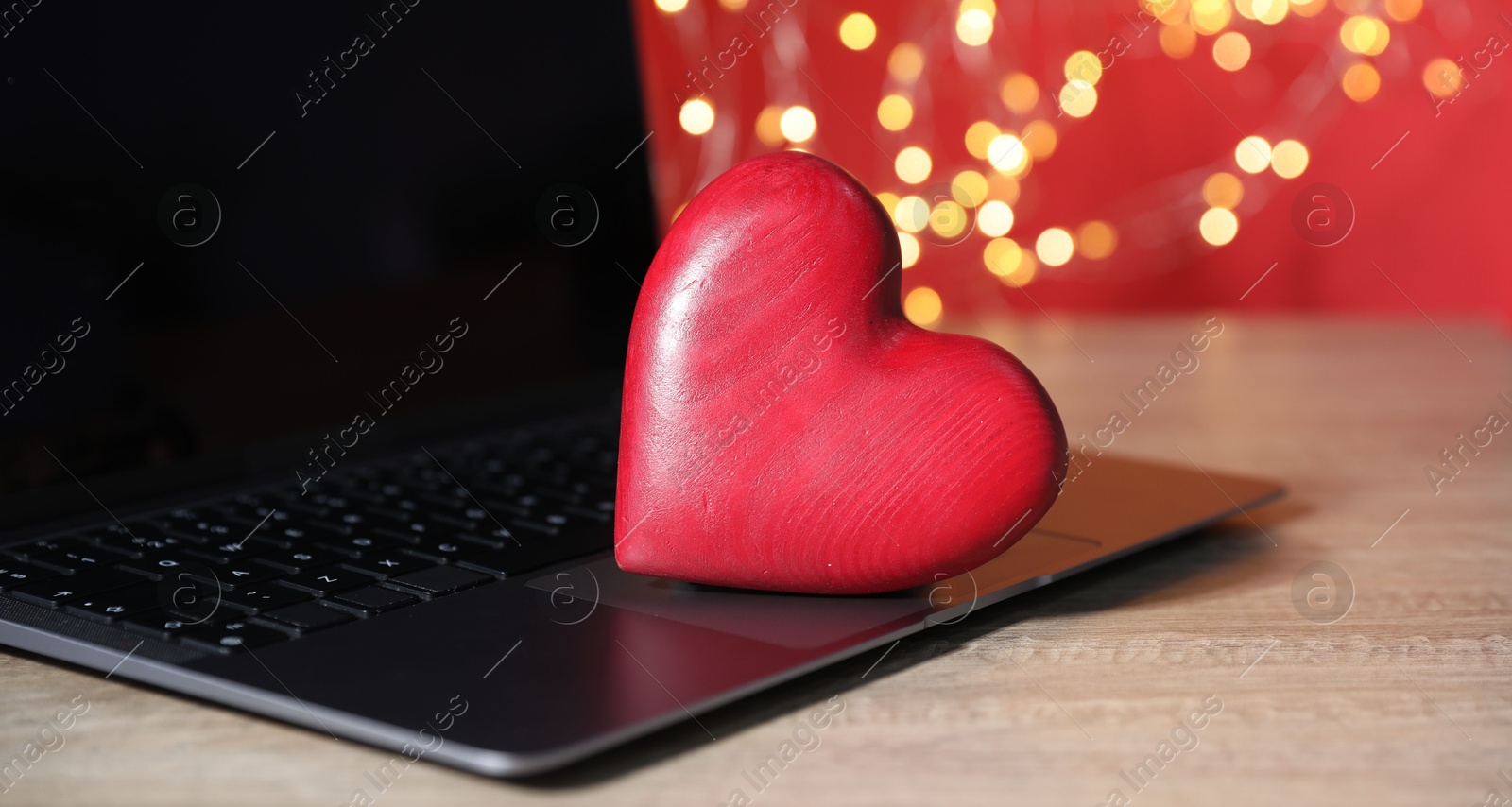 Photo of Online dating site. Red heart and laptop on wooden table against blurred lights, closeup. Bokeh effect