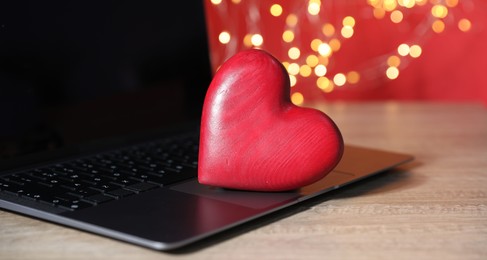 Photo of Online dating site. Red heart and laptop on wooden table against blurred lights, closeup. Bokeh effect