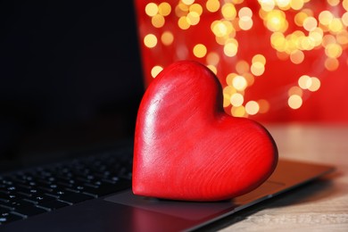 Photo of Online dating site. Red heart and laptop on wooden table against blurred lights, closeup. Bokeh effect