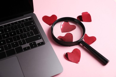 Photo of Online dating site. Many red hearts, magnifying glass and laptop on pink background, closeup