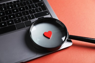 Photo of Online dating site. Laptop, heart and magnifying glass on red-brown background, closeup
