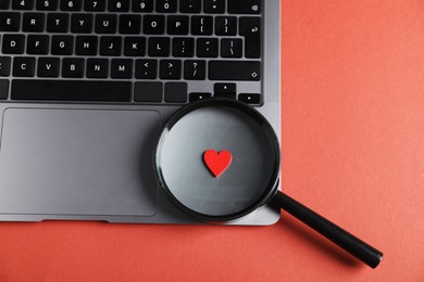 Photo of Online dating site. Laptop, heart and magnifying glass on red-brown background, top view