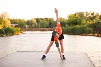 Photo of Young sportsman in headphones and armband doing morning exercise on pier near river