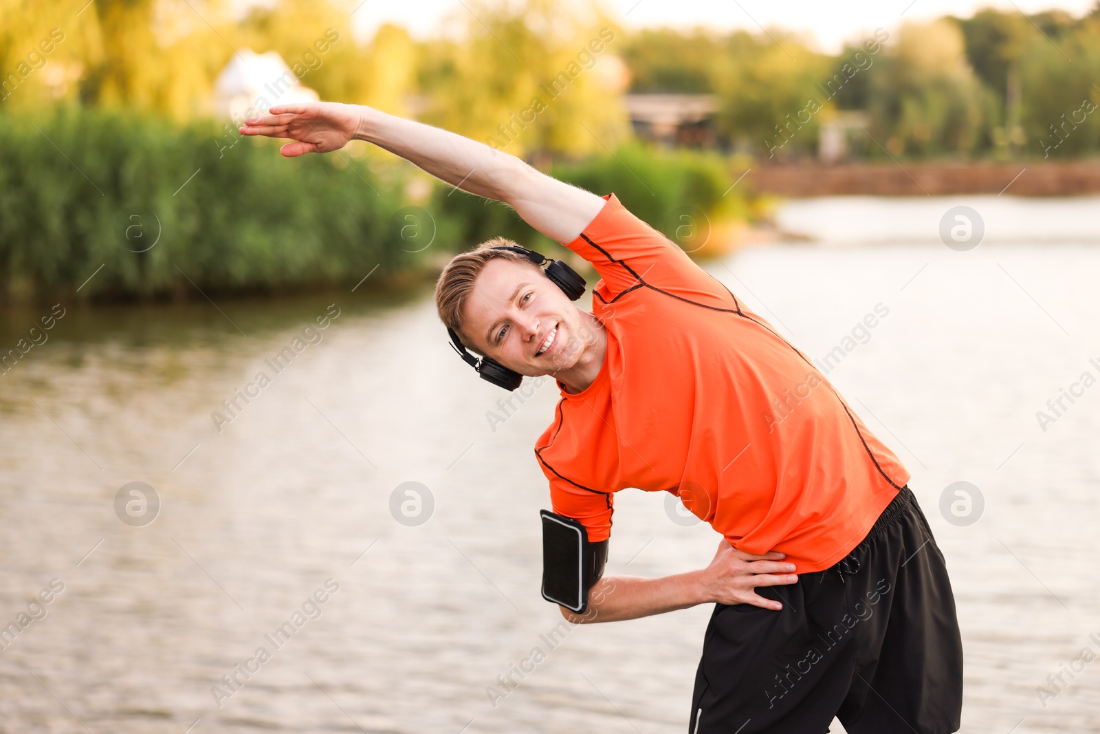 Photo of Smiling sportsman in headphones and armband doing morning exercise near river