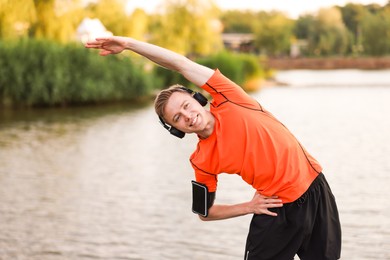 Photo of Smiling sportsman in headphones and armband doing morning exercise near river