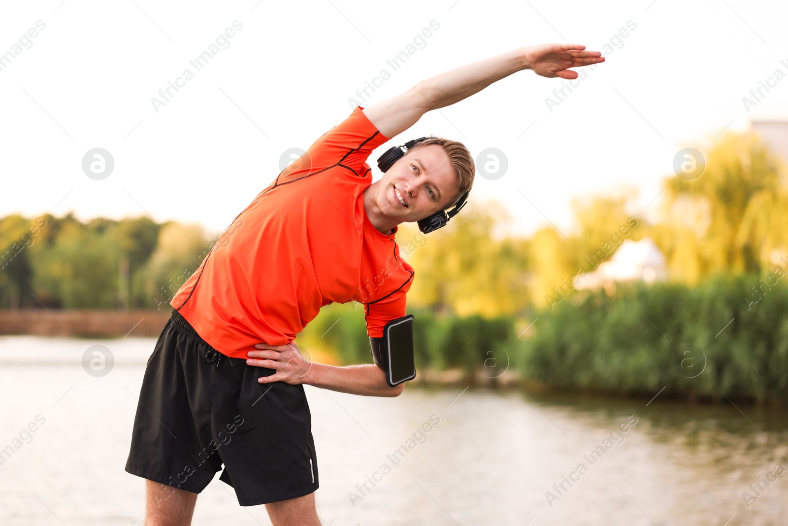 Photo of Smiling sportsman in headphones and armband doing morning exercise near river