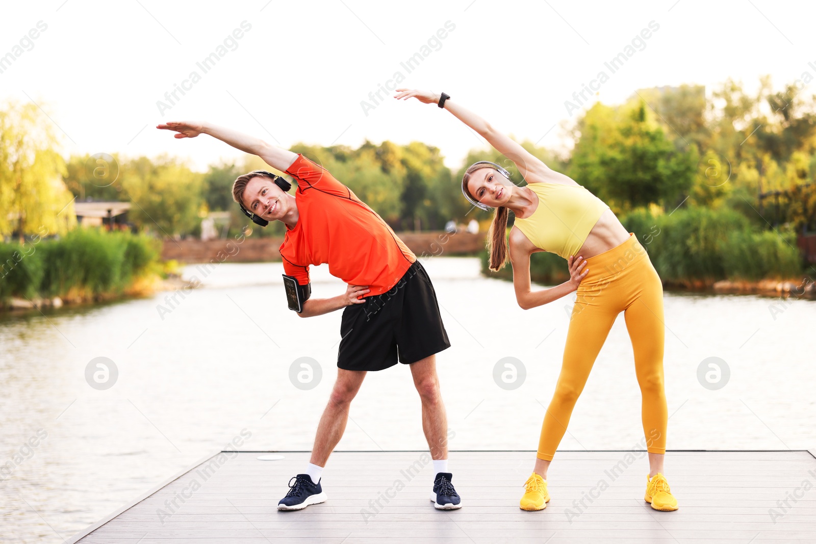 Photo of Smiling couple in headphones doing morning exercise on pier near river
