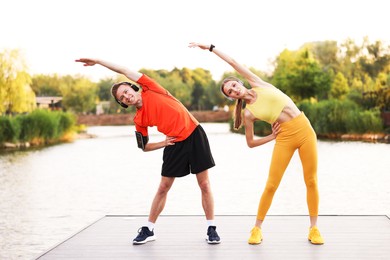 Photo of Smiling couple in headphones doing morning exercise on pier near river