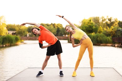 Photo of Smiling couple in headphones doing morning exercise on pier near river