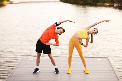 Photo of Smiling couple in headphones doing morning exercise on pier near river