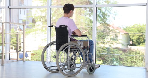 Photo of Young man in modern wheelchair near window indoors, back view