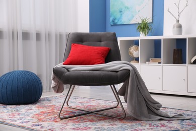 Photo of Comfortable grey armchair with red pillow and blanket in living room. Interior design