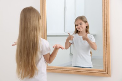 Image of Confused little girl in front of mirror where her reflection showing thumbs up indoors. Mental health