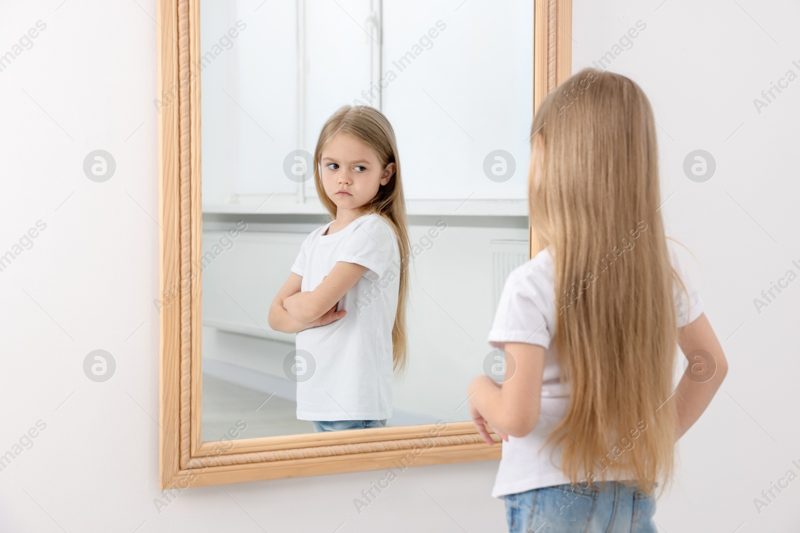 Image of Little girl looking at upset reflection of herself in mirror indoors. Mental health