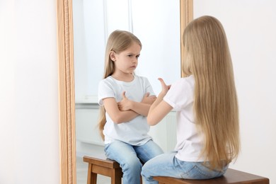 Image of Little girl showing thumbs up in front of mirror while her reflection demonstrating offended face expression indoors. Suppressed emotions