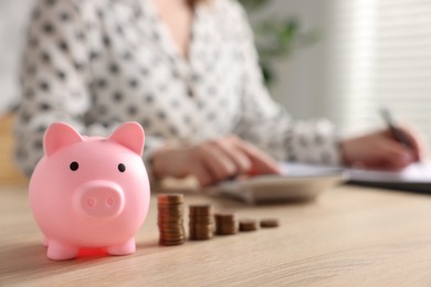 Photo of Piggybank, stacked coins and woman planning budget at wooden table, selective focus