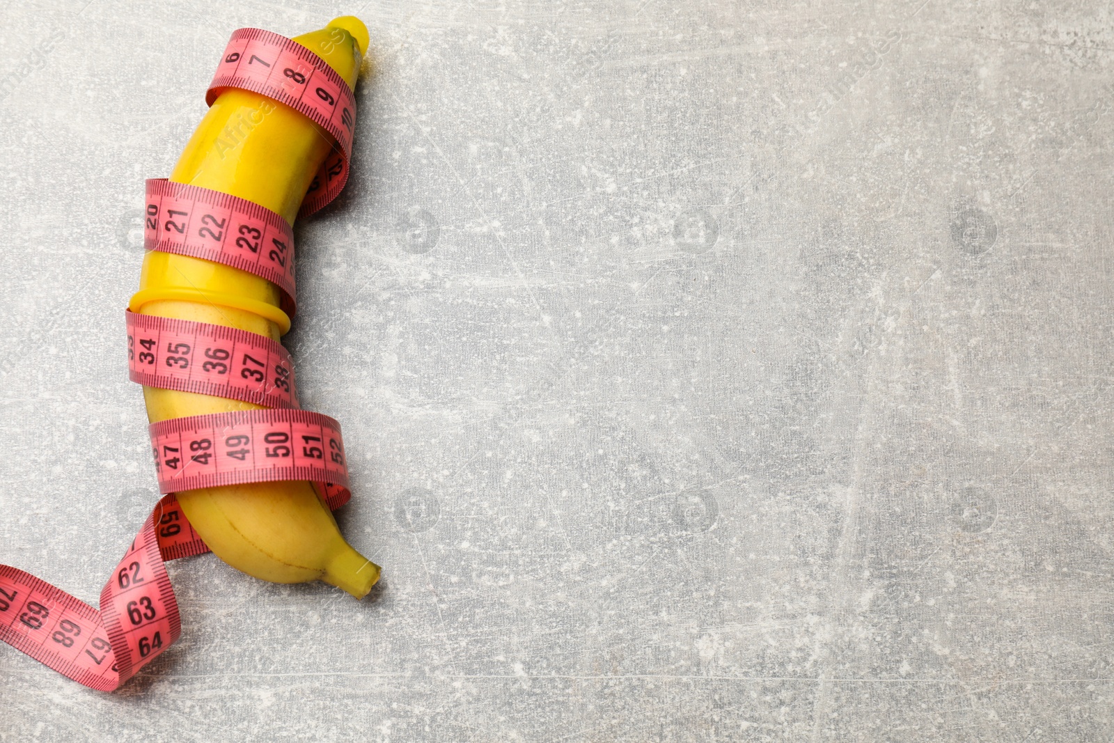 Photo of Sex education. Banana with condom and measuring tape on grey table, top view. Space for text