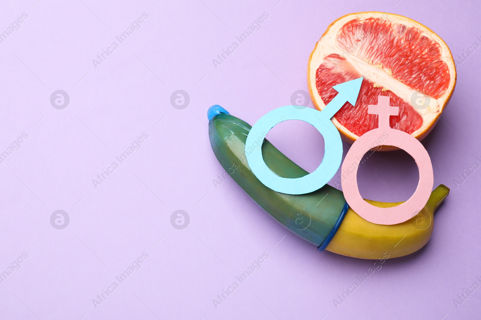 Photo of Sex education. Banana with condom, half of grapefruit and gender symbols on violet background, flat lay. Space for text