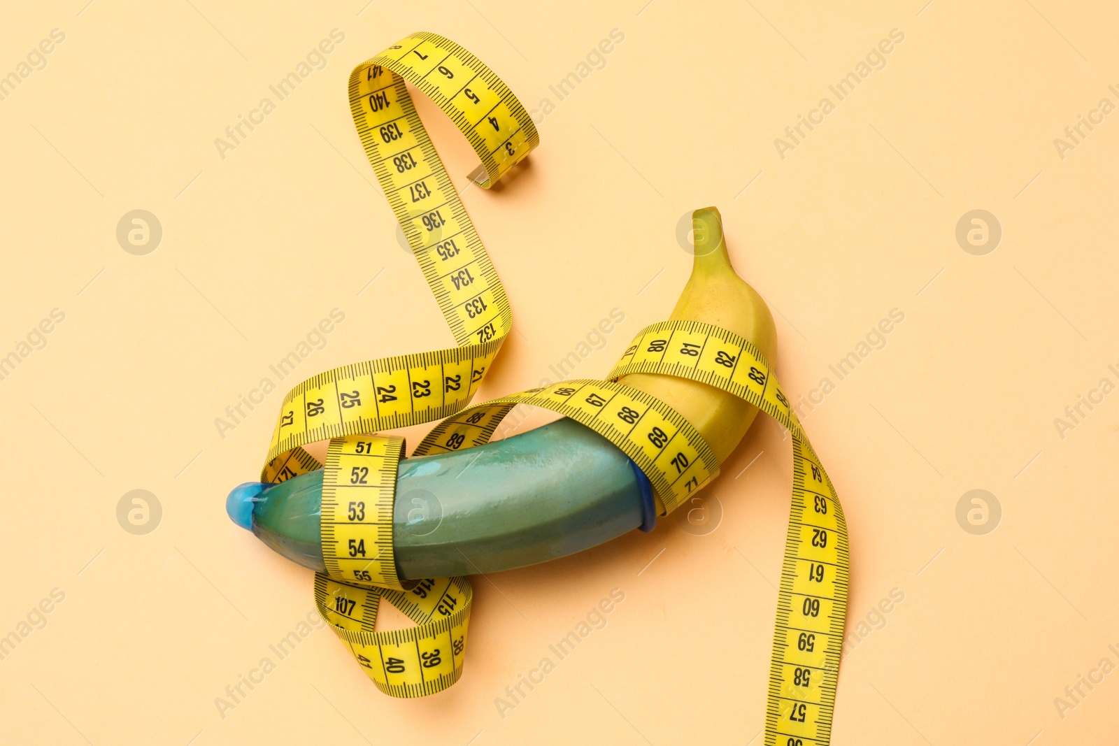 Photo of Sex education. Banana with condom and measuring tape on beige background, top view